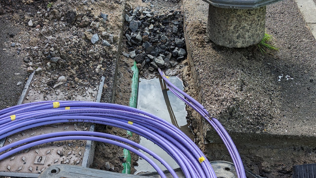 Purple fibre-optic cables laid in an excavated roadside trench during broadband network installation, with conduits and construction materials visible around the work area.